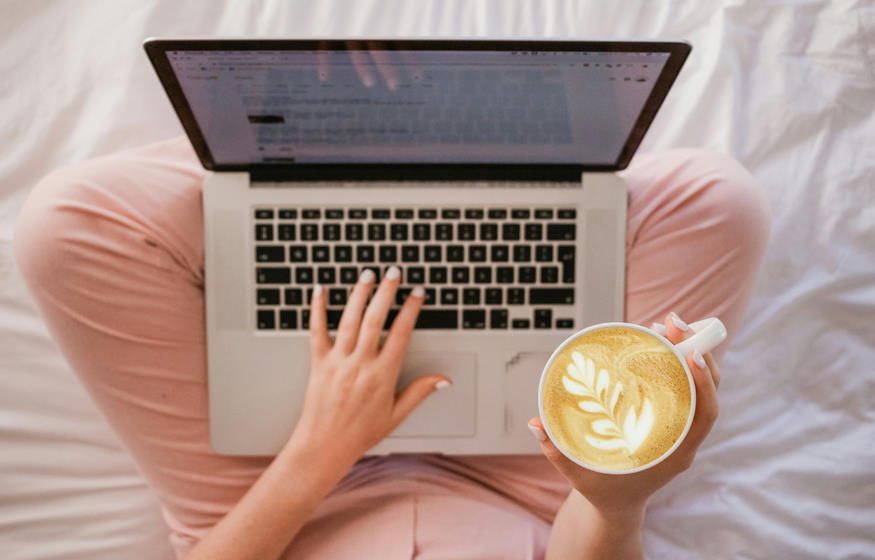 Une femme utilisant un MacBook Pro assise en tailleur sur son lit avec une tasse de café à la main, vue du dessus