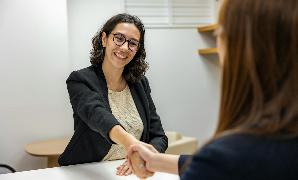 Une femme, brune avec des lunettes, serre la main de son interlocutrice après un entretient d'embauche réussi. Elle sourit.